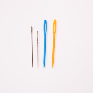 May include: Four sewing needles of varying sizes and colors are arranged on a white background. Two are metal, one is blue, and one is yellow. The needles are used for threading yarn or thread through fabric.