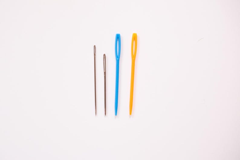 May include: Four sewing needles of varying sizes and colors are arranged on a white background. Two are metal, one is blue, and one is yellow. The needles are used for threading yarn or thread through fabric.