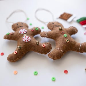 May include: Two brown felt gingerbread man ornaments with white string hangers. Each has a smiling face, candy accents, and colorful sequin buttons. Gold beads and colorful sequins are scattered around.