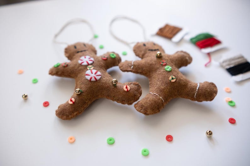 May include: Two brown felt gingerbread man ornaments with white string hangers. Each has a smiling face, candy accents, and colorful sequin buttons. Gold beads and colorful sequins are scattered around.