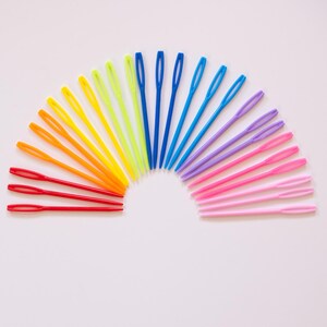 May include: A collection of colorful plastic yarn needles arranged in a semi-circle. The needles are in a rainbow of colors, including red, orange, yellow, green, blue, purple, and pink. The needles are on a white background.