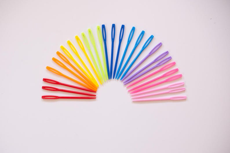 May include: A collection of colorful plastic yarn needles arranged in a semi-circle. The needles are in a rainbow of colors, including red, orange, yellow, green, blue, purple, and pink. The needles are on a white background.