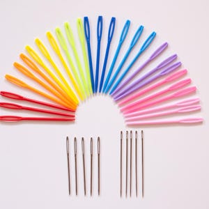 May include: A collection of colorful plastic yarn needles arranged in a semi-circle, with a rainbow of colors including red, orange, yellow, green, blue, purple, and pink. Below are several metal needles.