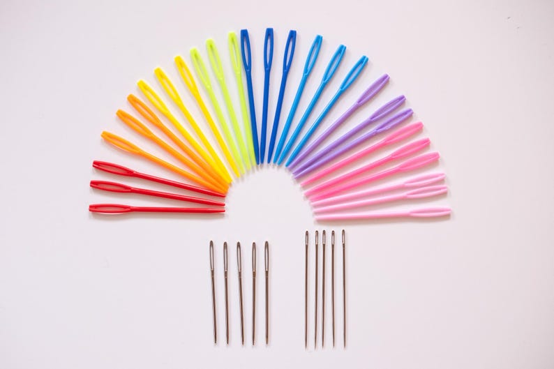 May include: A collection of colorful plastic yarn needles arranged in a semi-circle, with a rainbow of colors including red, orange, yellow, green, blue, purple, and pink. Below are several metal needles.