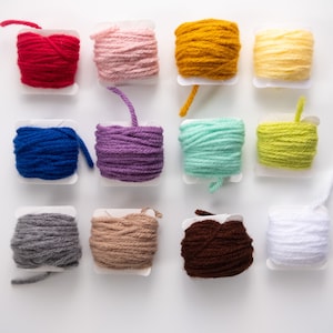 May include: Twelve balls of yarn in various colors, including red, pink, yellow, blue, purple, green, gray, brown, and white, are arranged in a grid pattern on a white surface.