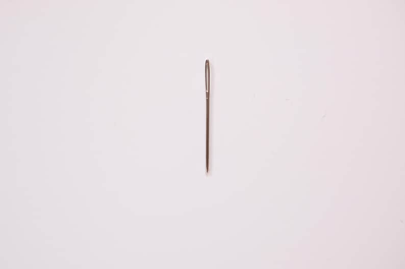 May include: A single, silver sewing needle with a small eye and a sharp point, centered on a white background. The needle is a common tool used for hand sewing and crafting projects.