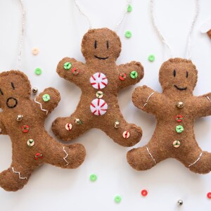 May include: Three brown felt gingerbread man ornaments with white stitching, button and sequin details, and white string hangers. One has a surprised expression, one has peppermint candies, and one has a smiling face. Christmas decor.