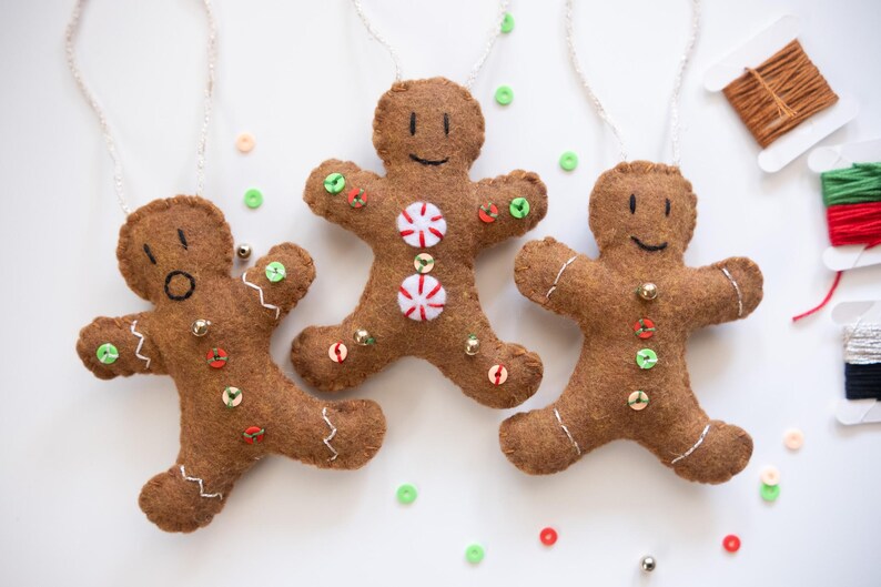 May include: Three brown felt gingerbread man ornaments with white stitching, button and sequin details, and white string hangers. One has a surprised expression, one has peppermint candies, and one has a smiling face. Christmas decor.