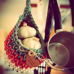 May include: A colourful, handmade crochet basket in red, green, and cream, suspended from a hook, contains several heads of garlic. A wooden heart-shaped tag reading "Handmade with Love" is attached, along with a small button with a heart design.