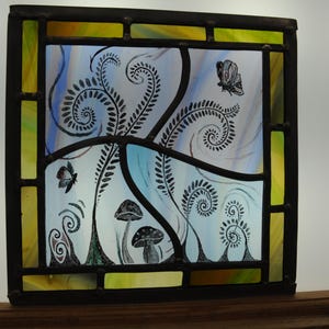 May include: A square stained glass panel framed in dark metal, featuring a light blue and yellow background. The glass displays black line art of two mushrooms, several swirling fern-like fronds, and two butterflies in a nature-inspired scene.