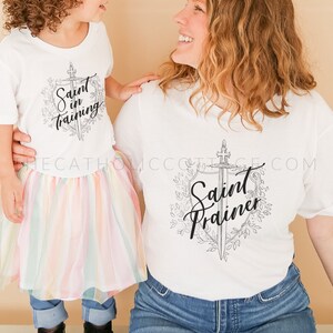 May include: A white t-shirt with the text "Saint in training" and a sword graphic. A second white t-shirt with the text "Saint Trainer" and a sword graphic surrounded by leaves. The shirts are worn by a child and an adult.