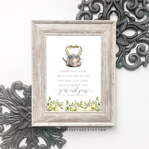 May include: A framed print featuring a watercolour illustration of a teapot and foliage. The text reads "KNOW THAT EVEN WHEN YOU ARE IN THE KITCHEN, OUR LORD MOVES AMIDST THE pots and pans." The frame is light grey, and the background is white.