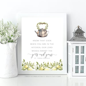 Puede incluir: Una impresión enmarcada blanca con una ilustración de acuarela de una tetera con una corona de limón. El texto dice "Sepa que incluso cuando está en la cocina, nuestro Señor se mueve en medio de las ollas y las sartenes. Santa Teresa de Ávila".