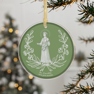 May include: Round Christmas ornament with a green background featuring a white illustration of St. Cecilia holding a musical instrument. The ornament has a gold ribbon for hanging and the text "St. Cecilia ora pro nobis".