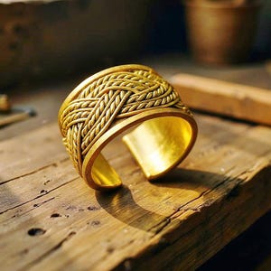 Puede incluir: Un brazalete dorado con un diseño trenzado. El brazalete abierto se encuentra sobre una superficie de madera. El intrincado diseño y el cálido color dorado se destacan. El ancho del brazalete es de aproximadamente 4 cm.