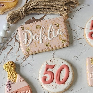 May include: A set of decorated sugar cookies with pink icing and gold accents. One cookie is decorated with the name "Dahlia", another with the number "50", and a third with a champagne bottle design.