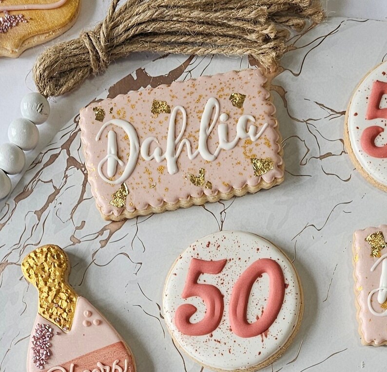 May include: A set of decorated sugar cookies with pink icing and gold accents. One cookie is decorated with the name "Dahlia", another with the number "50", and a third with a champagne bottle design.