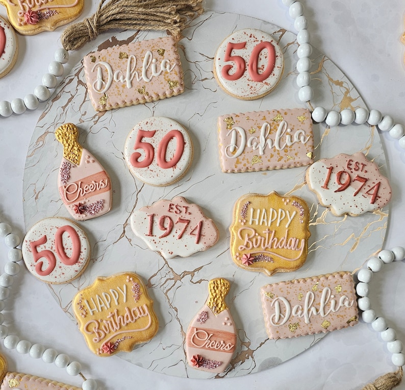 May include: A collection of decorated sugar cookies with various designs and text, including "Happy Birthday", "Cheers", "Dahlia", "50", and "EST. 1974".