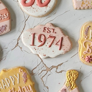 May include: A set of decorated sugar cookies with pink and white frosting. One cookie has the text "EST. 1974" in pink lettering. Another cookie has the text "Happy Birthday" in white lettering.