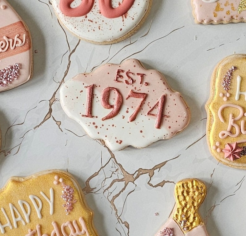 May include: A set of decorated sugar cookies with pink and white frosting. One cookie has the text "EST. 1974" in pink lettering. Another cookie has the text "Happy Birthday" in white lettering.