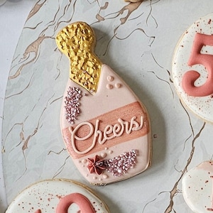 May include: A pink and gold decorated sugar cookie shaped like a champagne bottle with the word "Cheers" written on it.