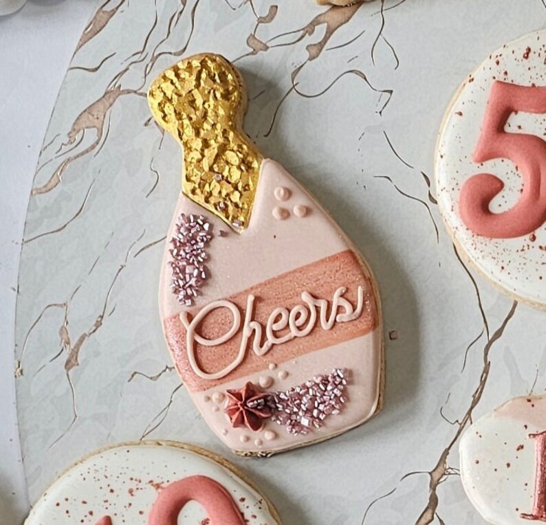 May include: A pink and gold decorated sugar cookie shaped like a champagne bottle with the word "Cheers" written on it.