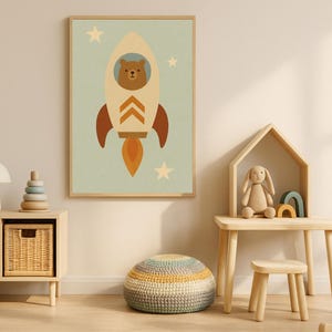 May include: A framed art print depicting a bear in a rocket ship against a light blue backdrop with stars. The rocket is cream with brown and orange details. The artwork is displayed in a child's room with other toys and furniture.