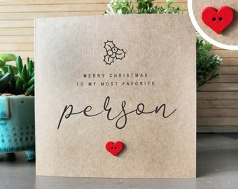 May include: A brown card with a black holly sprig and the text "Merry Christmas to my most favourite person" in black script. There is a red heart-shaped button on the card.
