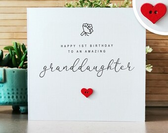 Op de afbeelding: Witte kaart met zwarte tekst die "Happy 1st Birthday to an amazing granddaughter" luidt. In de linkerbovenhoek staat een kleine zwart-witte illustratie van een boeket bloemen. Op de kaart zitten twee rode hartvormige knopen.