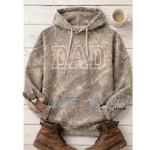 Puede incluir: Una sudadera con capucha de estampado de camuflaje que presenta la palabra DAD en grandes letras blancas en el pecho. La prenda incluye una capucha con cordón, un bolsillo tipo canguro y un pequeño gráfico de una llama marrón abajo a la izquierda.