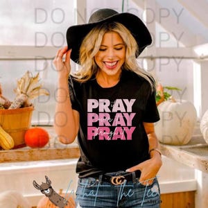 Puede incluir: Camiseta negra con el texto "PRAY it over PRAY through PRAY" en rosa y blanco. La camiseta se usa con vaqueros azules y un sombrero negro. La imagen incluye calabazas y un gráfico de llama.