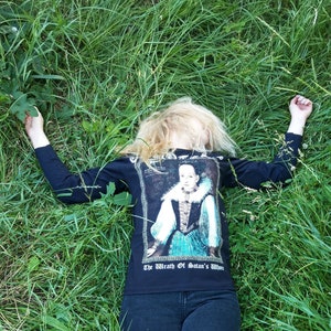 May include: A black long-sleeved shirt with a graphic print of a woman in a blue and white dress. The text "The Wrath of Satan's Wife" is printed below the image.