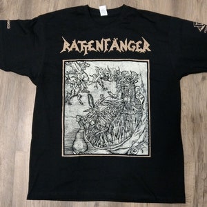 Op de afbeelding: Zwart t-shirt met het woord "RATTENFÄNGER" in bruine letters boven een gedetailleerde zwart-wit illustratie. De illustratie toont een complexe scène met figuren en een centrale structuur. De mouwen hebben tekst en een logo.