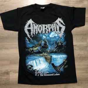 May include: Black t-shirt with a silver band logo for the band Amorphis. The graphic features a fantasy landscape with a waterfall and the text "Tales from the Thousand Lakes".