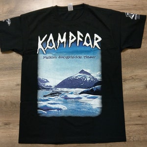 May include: Black t-shirt with a white graphic print of the band Kampfar. The graphic features the band's name in a stylized font and the text "Mellom Skogkledde Tårer". The graphic also includes a blue and white image of a mountain range and a body of water.