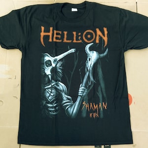 Puede incluir: Camiseta negra con un estampado gráfico de una criatura esquelética sosteniendo un bastón coronado con una calavera. El texto "Hellion" y "Shaman" está impreso en la camiseta en naranja.
