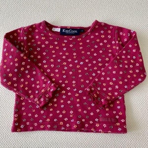 May include: A long-sleeved, burgundy shirt with a small floral print. The shirt has a ruffled trim on the sleeves and the KidCool Collections logo is visible on the front.