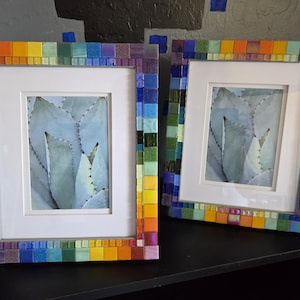 May include: Two rectangular picture frames featuring a vibrant mosaic border made of small, multi-colored square tiles in shades of red, orange, yellow, green, blue, and purple. Each frame contains a white mat and a print of a pale green agave plant.