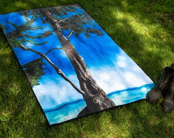 May include: A blue and green beach towel with a large tree and a blue sky printed on it. The towel is lying on green grass.