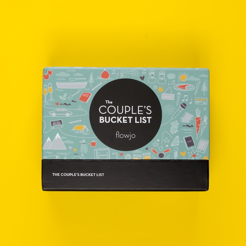 May include: A blue and white box with illustrations of a couple's bucket list. The box has the text "The Couple's Bucket List" and "flowjo" on it. The bottom of the box has the text "The Couple's Bucket List" in black.