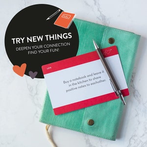 May include: A red and white card with the text "Buy a notebook and leave it in the kitchen to share positive notes to eachother." The card is on top of a teal notebook with a pen on top of the card. The card has two small hearts in the top right corner.