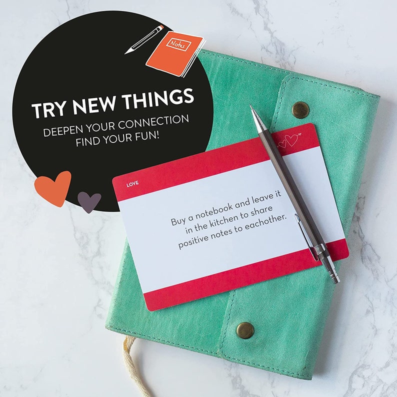 May include: A red and white card with the text "Buy a notebook and leave it in the kitchen to share positive notes to eachother." The card is on top of a teal notebook with a pen on top of the card. The card has two small hearts in the top right corner.