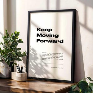 Puede incluir: Un póster motivacional enmarcado en negro con fondo blanco y el texto "Keep Moving Forward". El póster está colgado en una pared blanca con una planta en una maceta y lápices en un porta lápices sobre una mesa de madera.