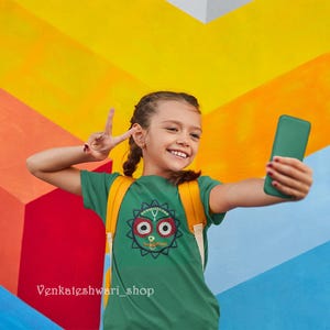 May include: A young person wearing a green t-shirt with a colorful graphic design of a face. The person is holding a green smartphone and making a peace sign with their hand. The background is a colorful wall with stripes of yellow, orange, blue, and red.