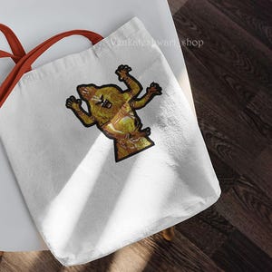 May include: A white tote bag with orange straps features a brown and yellow embroidered image of a creature with four arms and a long tail.
