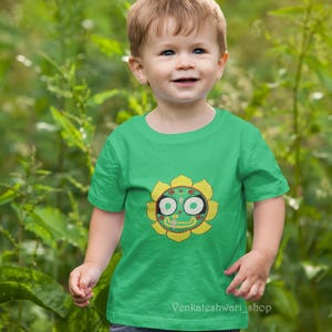 May include: A young child wearing a green t-shirt with a colorful sunflower design. The sunflower has a face with large eyes and a smiling mouth. The child is standing in front of a green background.