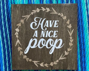 May include: A wooden sign with a gold-colored wreath and the text "Have a nice poop" in white lettering.
