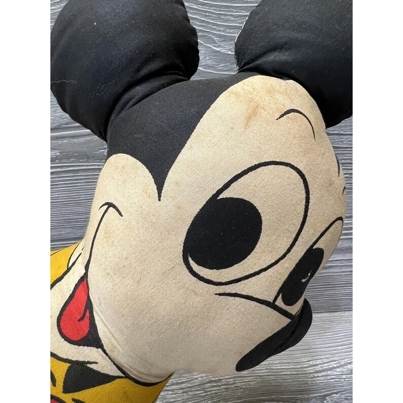 May include: Close-up of a vintage Mickey Mouse plush toy. The toy is made of fabric and has a black and white face with a red mouth. The toy is in good condition, but shows signs of wear and tear.