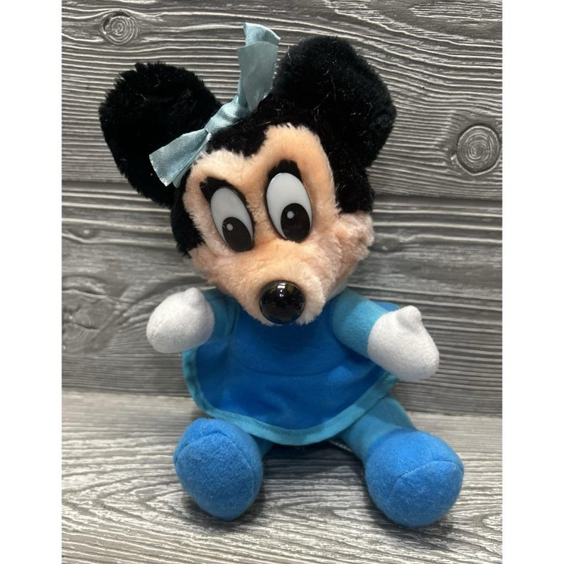 Pode incluir: Uma boneca de pel&uacute;cia da Minnie Mouse com orelhas pretas, um la&ccedil;o azul claro e um vestido azul. A boneca tem um rosto rosa, olhos grandes e luvas brancas. A boneca est&aacute; sentada.