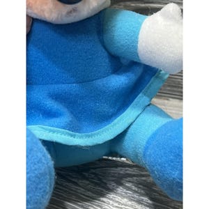 Pode incluir: Uma boneca de pel&uacute;cia com uma roupa azul e detalhes brancos. O corpo da boneca &eacute; principalmente azul, com detalhes em azul claro e branco nos bra&ccedil;os e na cabe&ccedil;a. A boneca &eacute; feita de material macio e felpudo.